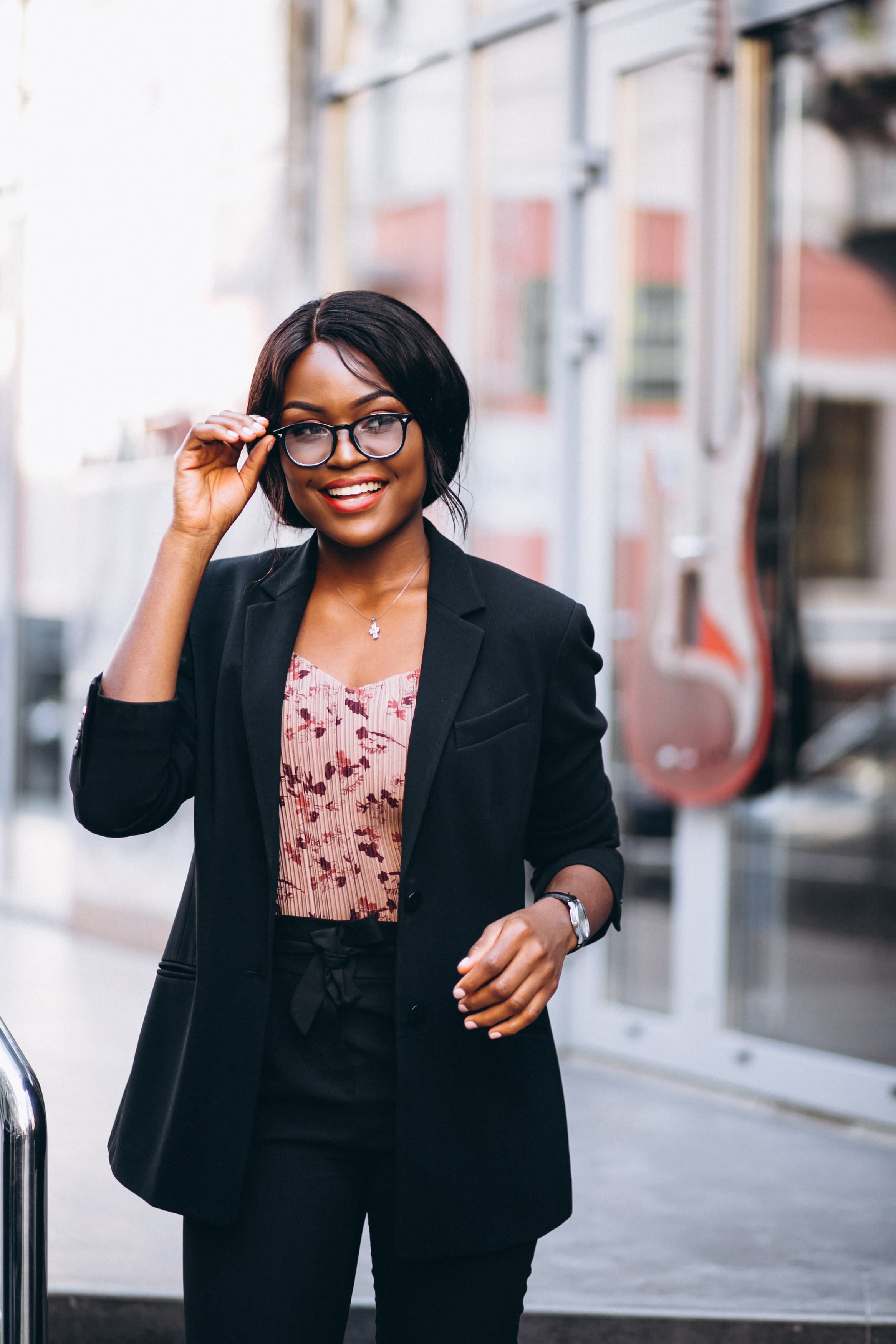 Smiling Black woman in a black blazer adjusts her glasses while standing outside a building.